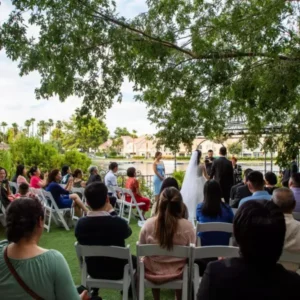 Lakeside wedding ceremony with guests seated and bride and groom exchanging vows by the water, surrounded by greenery and palm trees, capturing the romantic atmosphere of a Las Vegas lakeside venue.