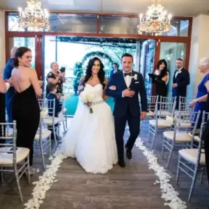 Bride in a white wedding gown walking down the aisle with her father, surrounded by guests in a lakefront wedding chapel with elegant chandeliers and floral decorations, showcasing the Lakeside Weddings & Events venue.
