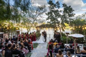 Outdoor wedding ceremony at Lakeside Weddings venue, featuring a couple exchanging vows, surrounded by lush greenery, a lake view, and illuminated by string lights, with guests seated on either side.