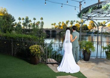 Couple in wedding attire standing by a lake, gazing at the water and palm trees, with a gazebo in the background, showcasing a romantic outdoor wedding setting in Las Vegas.