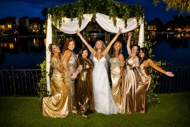 Bride celebrating with bridesmaids in gold dresses under a floral arch at a Best of Las Vegas 2026 lakeside wedding venue